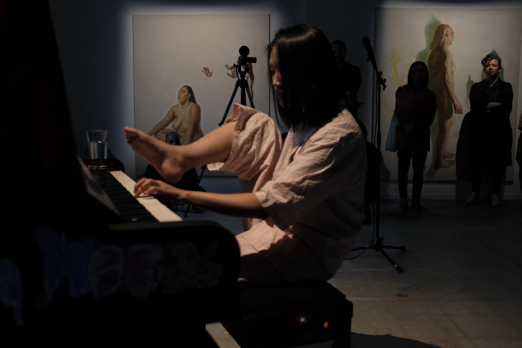 Fotografia della serie Kinderszenen di Double A Lab: un'artista asiatica seduta al pianoforte in una galleria d'arte, catturata in un momento di performance espressiva con opere d'arte contemporanea